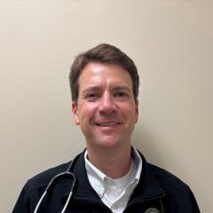 Man with short hair and stethoscope smiling against a light-colored wall.