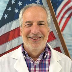 Smiling man in white lab coat and plaid shirt with American flag in background.
