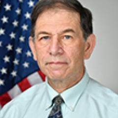 A man in a light blue shirt and tie stands in front of an American flag.