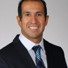 Man in black suit and blue tie smiling against a gray background.