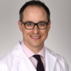 A smiling man in a white lab coat, pink shirt, and black tie.