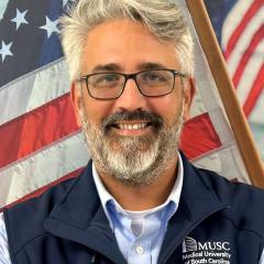A smiling man with a beard and glasses in front of an American flag.