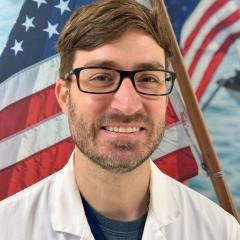 Man in white lab coat and glasses smiling in front of an American flag.