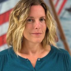 Woman with blonde hair and blue shirt in front of American flag.