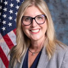 Woman with blonde hair wearing glasses and a gray blazer in front of an American flag.