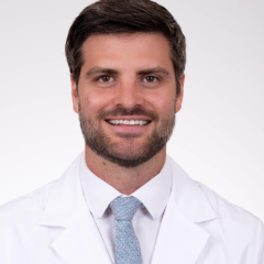 Man with short hair and beard in white lab coat and blue tie smiling.