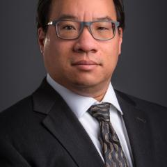 Man in black suit and tie, wearing glasses, against a black background.