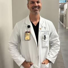 Smiling male doctor in white lab coat with name tag stands in hallway with hands in pockets.