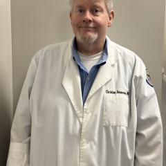 Man in white lab coat with blue shirt underneath, smiling against a plain wall.