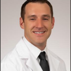 Male doctor in white lab coat and black tie smiling at camera.