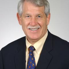 Portrait of a man with gray hair wearing a black suit and blue tie smiling.