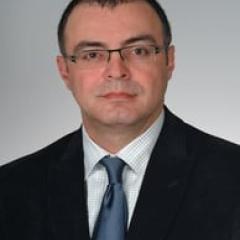 Man in black suit and blue tie with glasses, looking at camera.