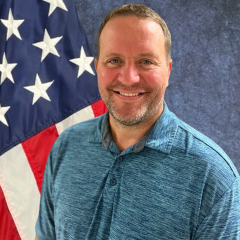 Man smiling in blue shirt in front of American flag.