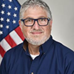 Man with gray hair, glasses, and blue shirt standing in front of an American flag.