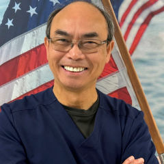 Man in blue shirt smiling with arms crossed in front of American flag.