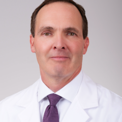 A man in a white lab coat and purple tie smiles for the camera.
