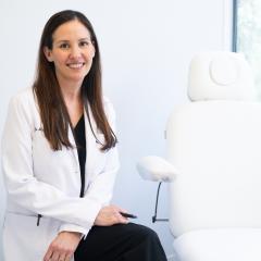 Woman in white lab coat smiling next to a medical chair.
