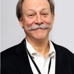 Portrait of an older man with gray hair, mustache, wearing a black blazer over a white shirt.