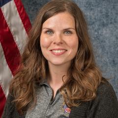 Portrait of smiling woman with long brown hair in front of American flag.