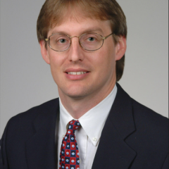 A man with glasses and a blue suit with a red and blue tie smiles.