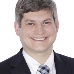 Portrait of a smiling man in a dark suit and blue plaid tie.