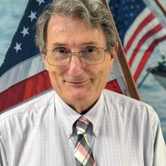 Man with gray hair, glasses, and plaid tie stands before two American flags.