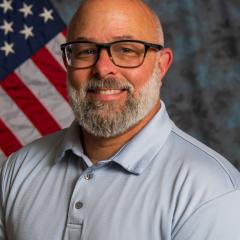Bald man with glasses and beard smiling, USA flag in background.