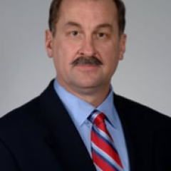 Man with mustache in black suit and blue shirt with red and white striped tie.