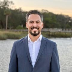 Man in a blue suit and white shirt smiles by a waterfront.