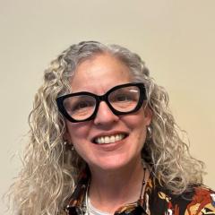 Portrait of a woman with curly gray hair and glasses, smiling.