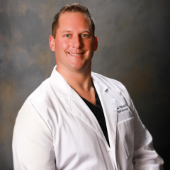 Smiling man in white lab coat and black shirt poses against gray background.
