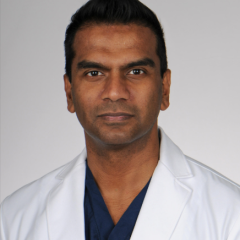 A man in a white lab coat and blue shirt stands against a white background.