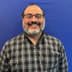 A man with glasses and a beard smiles against a blue backdrop.