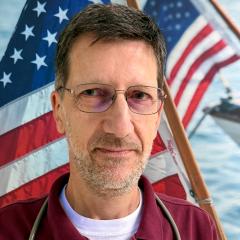 A man with a beard and glasses wearing a maroon shirt and stethoscope in front of an American flag.