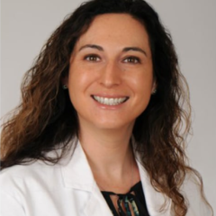 A smiling woman with curly hair in a white lab coat and black inner shirt.