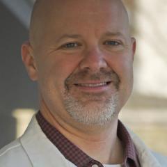 Photograph of a smiling bald man in a white coat and red shirt.