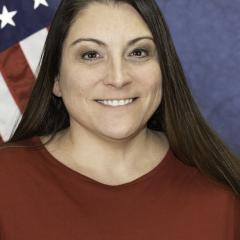 Woman with long brown hair wearing a red shirt smiling in front of an American flag.