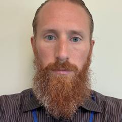 Man with red beard and blue lanyard, looking at camera.