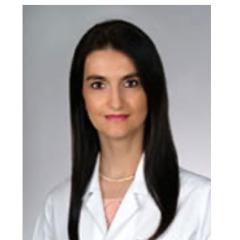 A smiling woman with long black hair wearing a white lab coat and necklace.