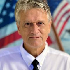 Portrait of a man in a white shirt and black tie, standing in front of an American flag.