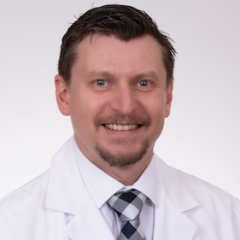 A smiling man in a white lab coat and checkered tie.