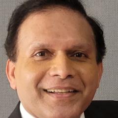 A man with short black hair, wearing a black suit and white shirt, smiling.