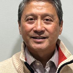Portrait of a man with short grey hair, wearing a beige sweater, smiling.