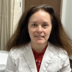 Woman in white lab coat over red shirt smiling in front of curtain.