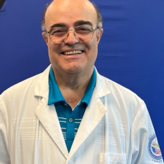 Man in white lab coat and blue polo shirt smiling against blue background.