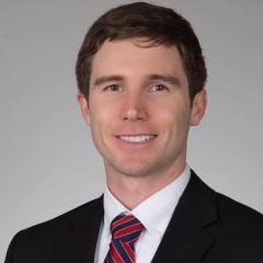 A man in a black suit and red, white, and blue striped tie smiles.