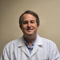 Man in a white lab coat with blue shirt underneath smiling at the camera.