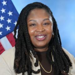 Headshot of a smiling woman with dreadlocks in front of an American flag.