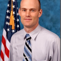 Man with gray shirt, blue tie, and American flag in background.