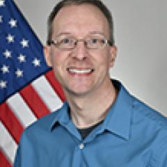 Man in blue shirt smiling, with American flag in background.